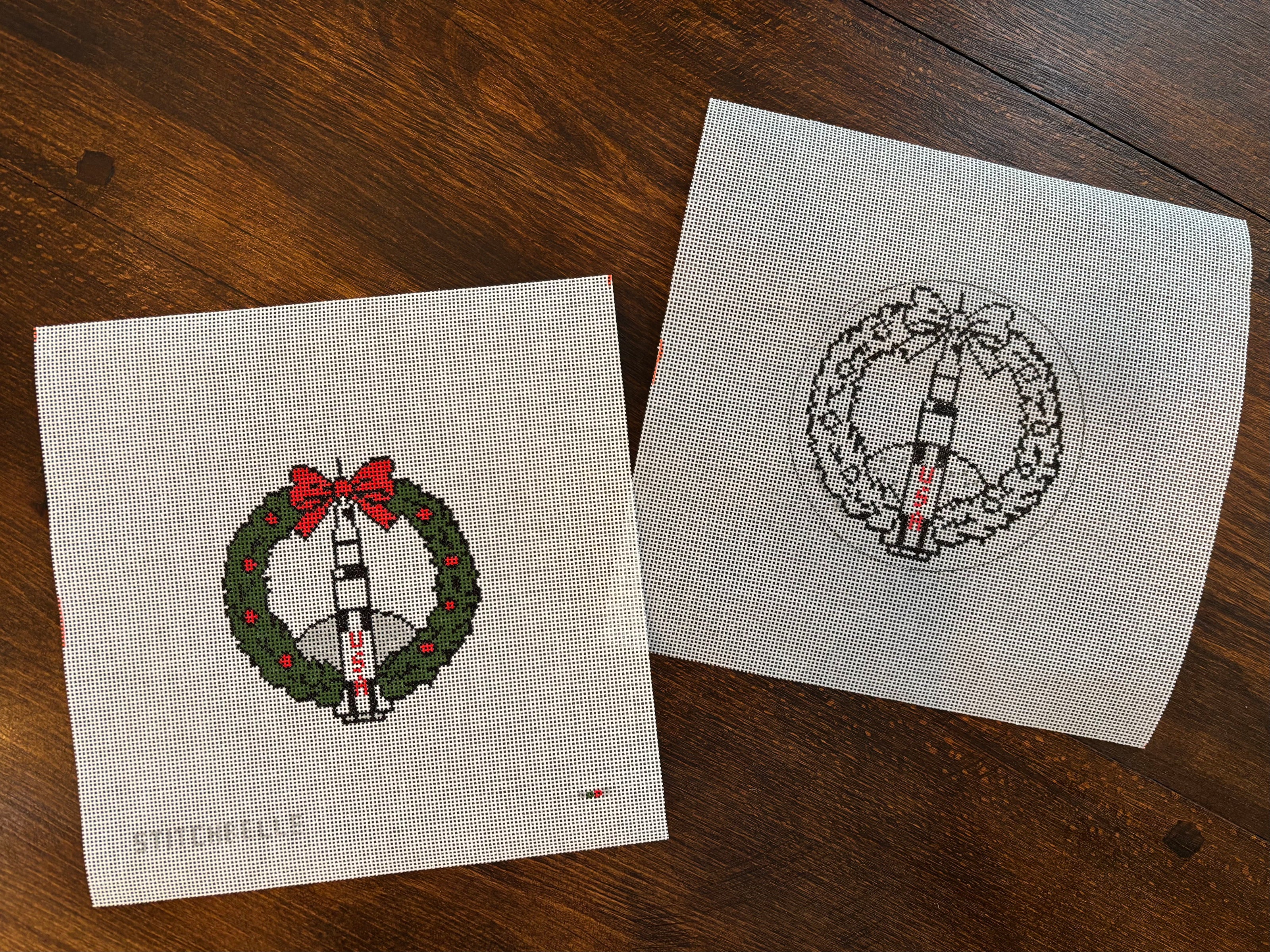 Two cross-stitched wreath patterns on fabric squares, one with a red bow and the other with a black and white design, on a wooden surface.